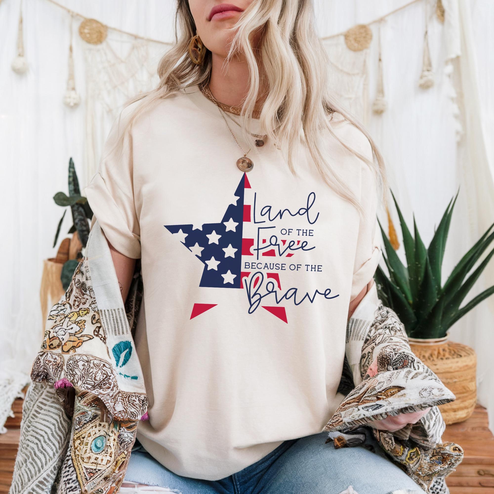 A woman wearing a white t-shirt with a patriotic design and text, sitting in front of a beige curtain with plants and a string of lights.