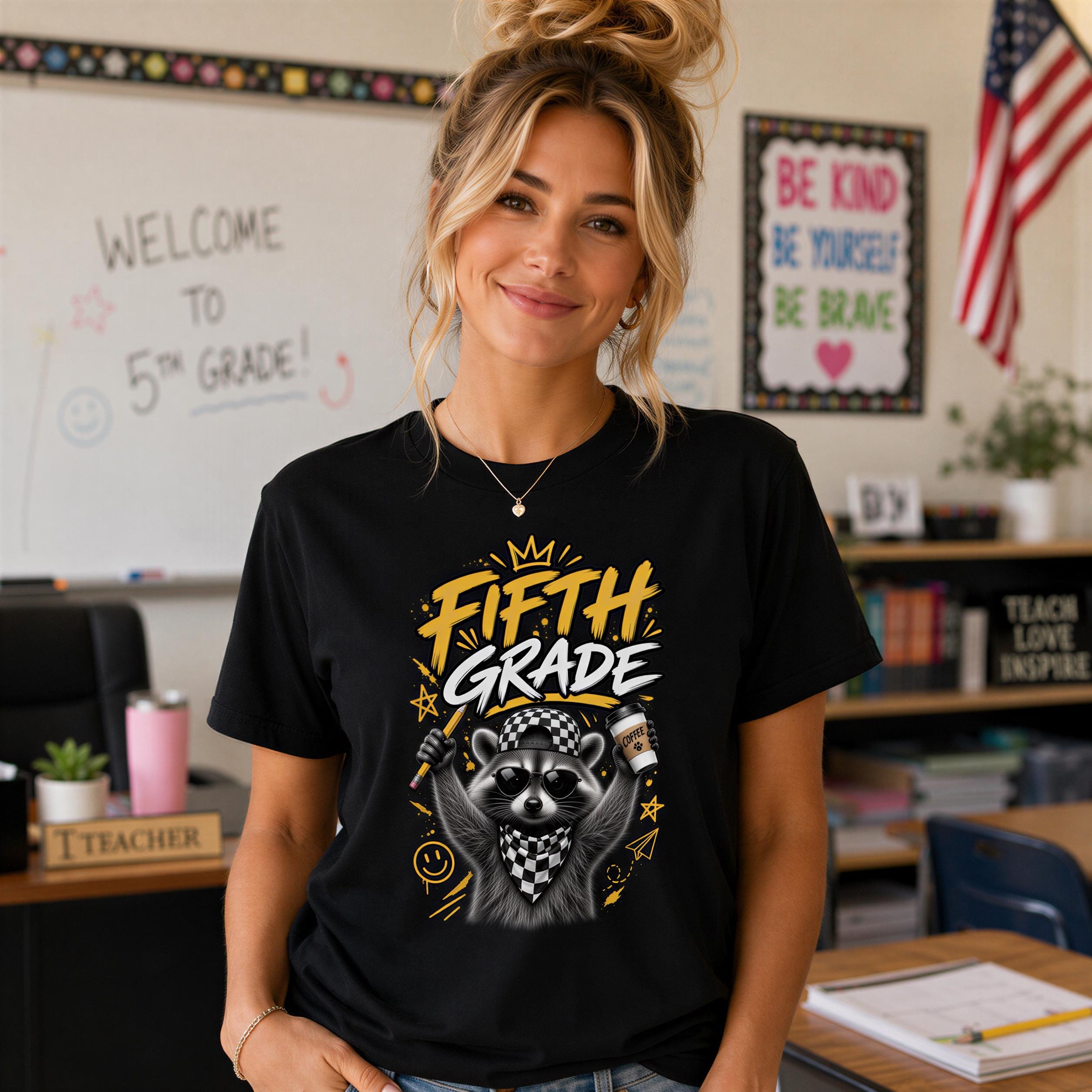 A smiling woman wearing a black t-shirt with a graphic of a raccoon and the text "Fifth Grade" stands in a classroom with a whiteboard and American flag in the background.