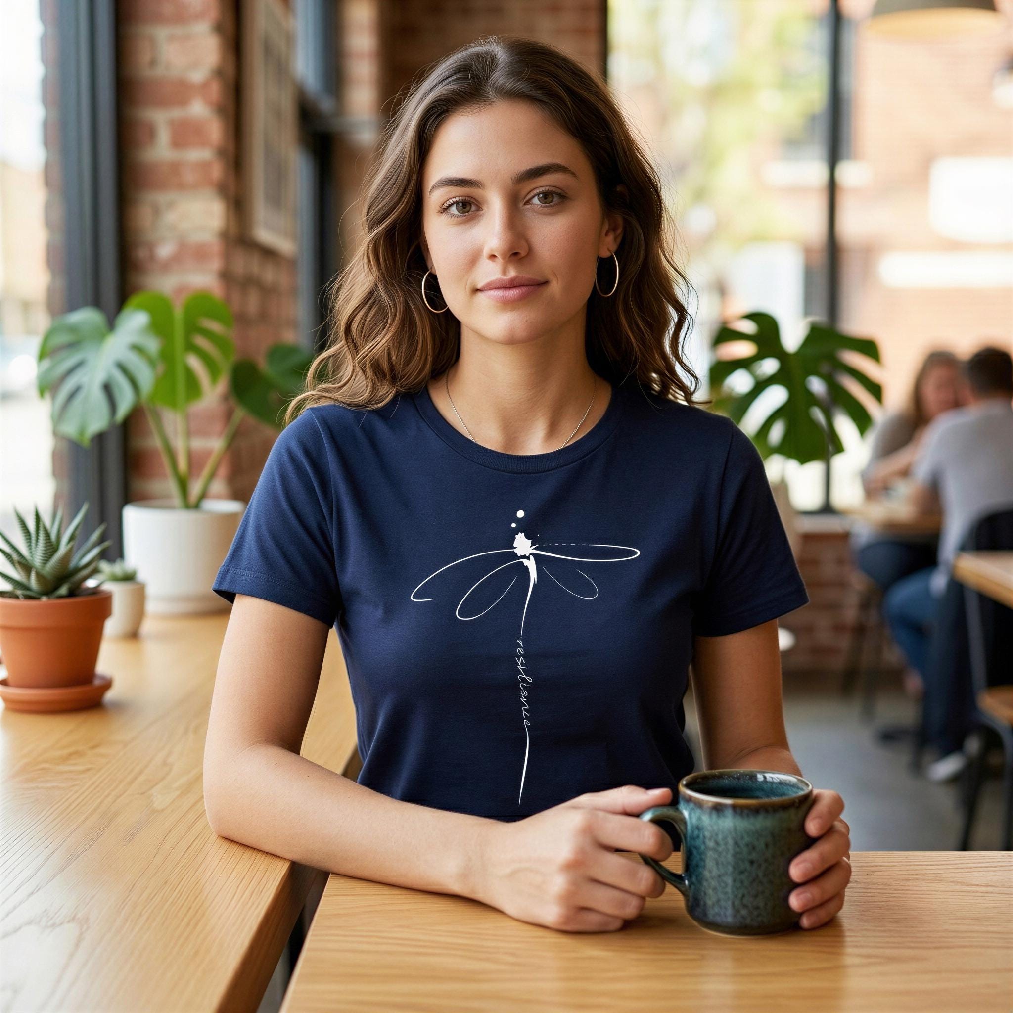 A young woman wearing a navy blue t-shirt with a white dragonfly design is sitting at a table in a cafe, holding a green mug.