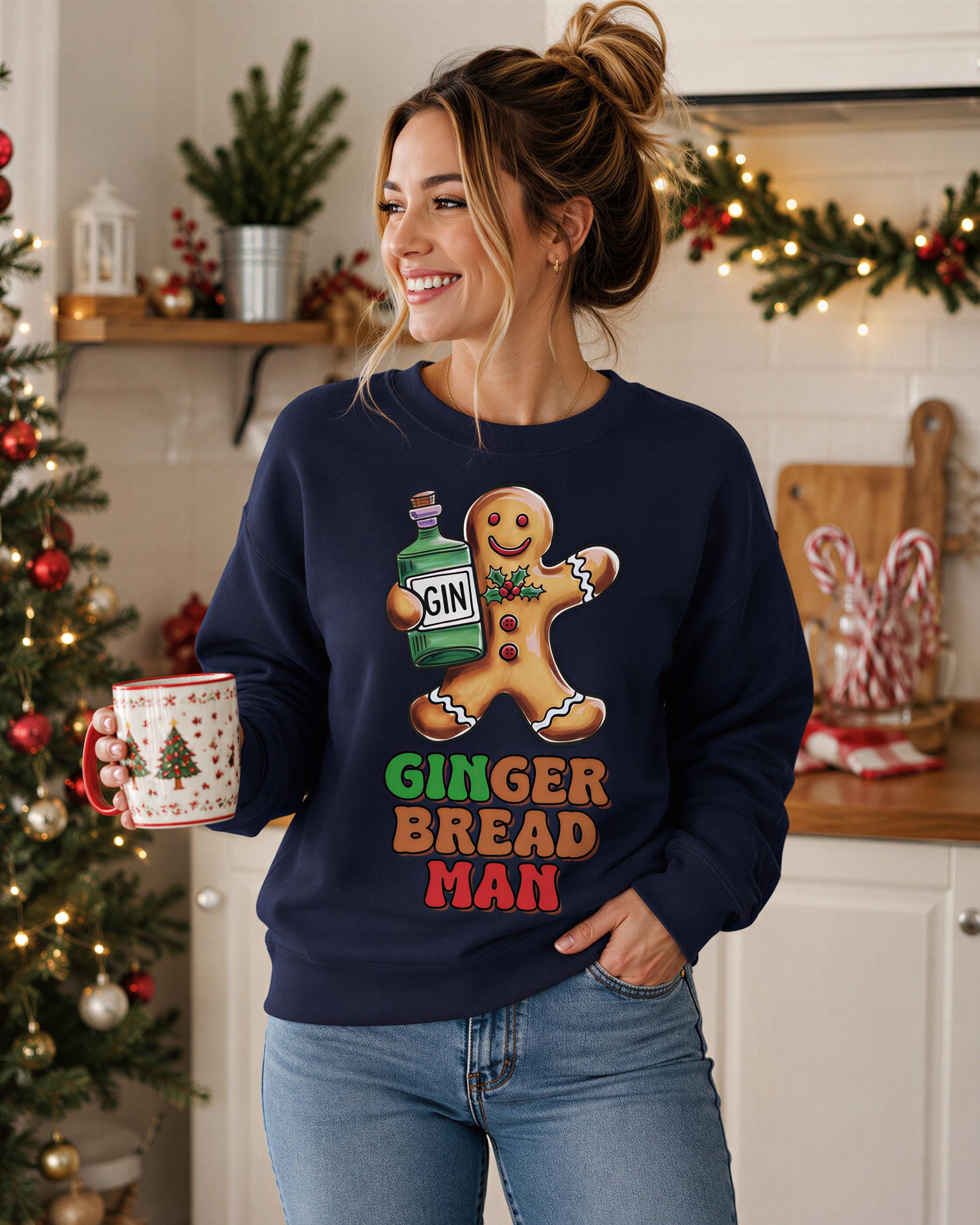 A woman wearing a navy blue sweatshirt with a graphic of a gingerbread man holding a bottle of gin, standing in a kitchen with Christmas decorations visible in the background.