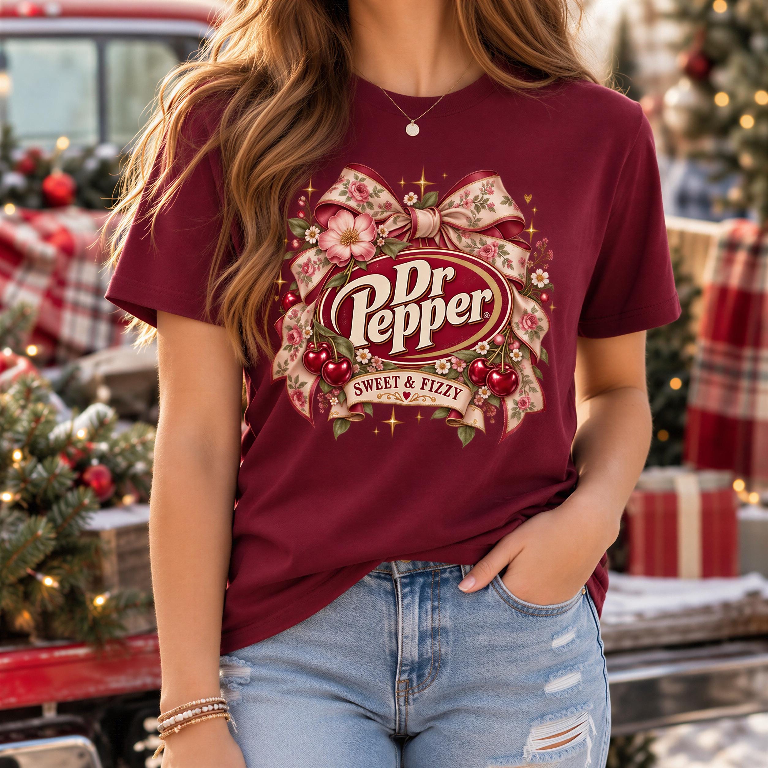 A woman wearing a burgundy t-shirt with a graphic of Dr Pepper and a bow, standing in front of a Christmas tree.