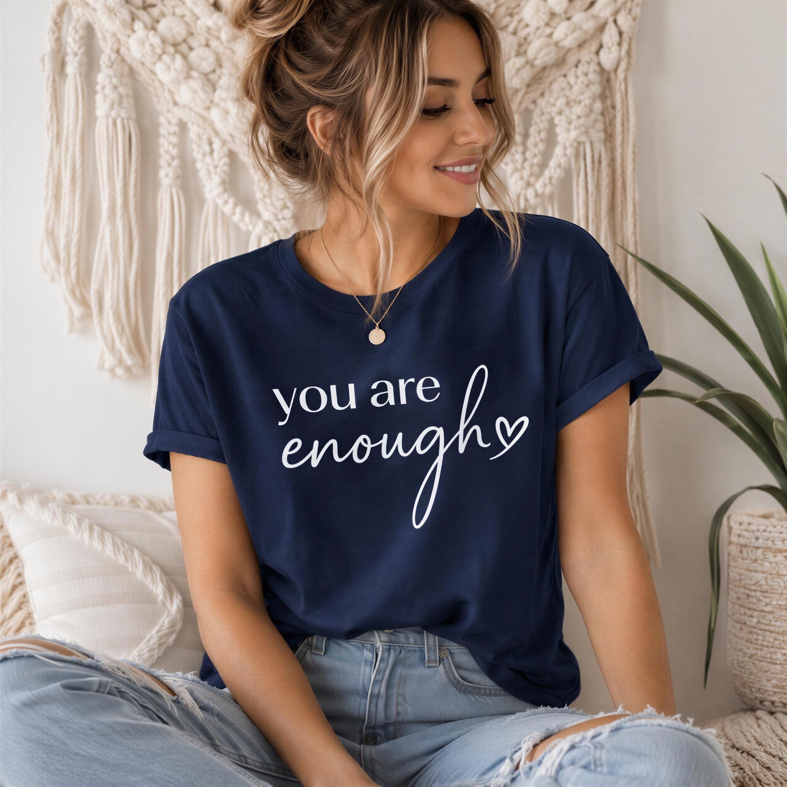 A woman wearing a navy blue t-shirt with the text "you are enough" is sitting on a couch, smiling and looking at the camera.