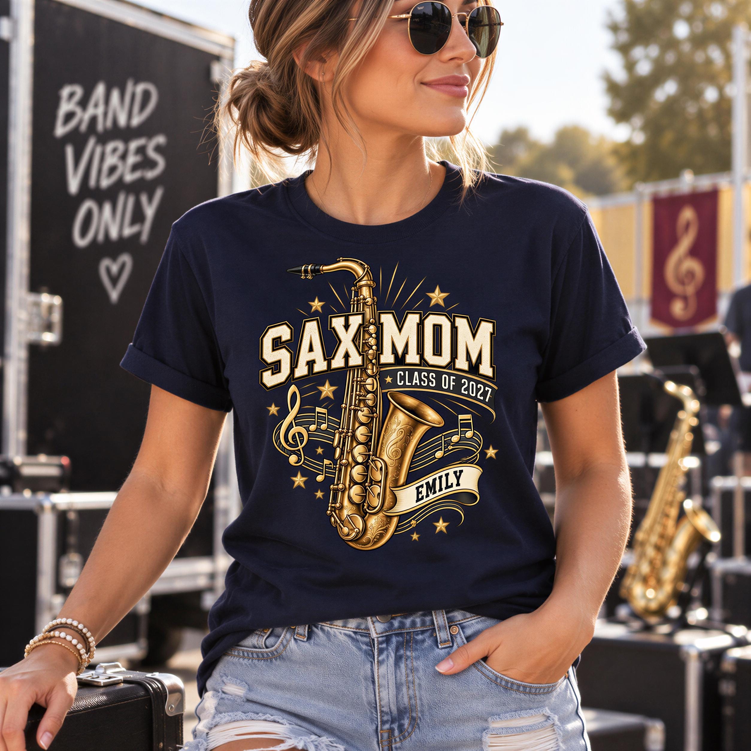 A woman wearing a navy blue t-shirt with a graphic design of a saxophone and the text "SAXMOM CLASS OF 2027 EMILY" is standing in front of a banner that says "BAND VIBES ONLY".