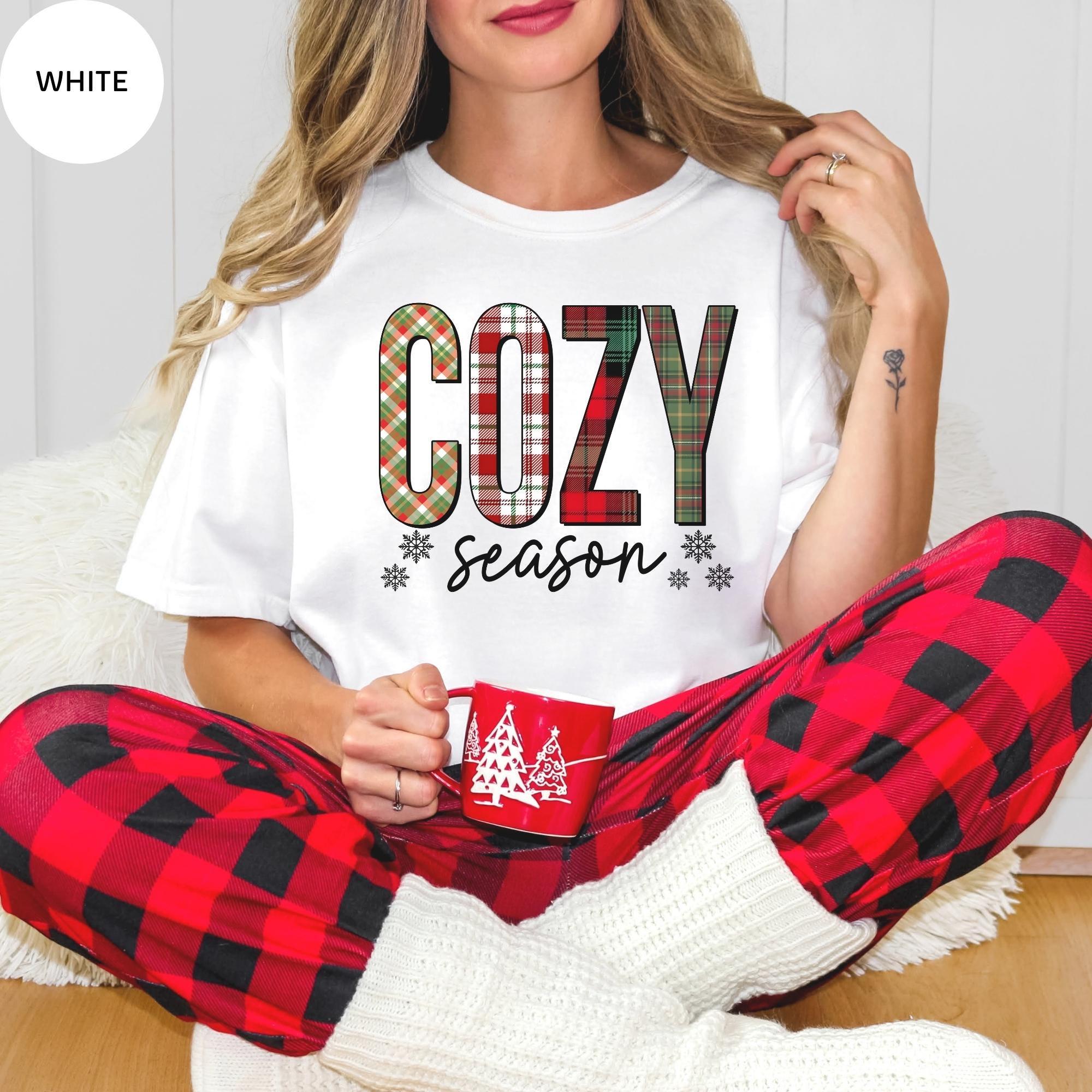 A woman wearing a white t-shirt with the word "COZY" and red plaid pajama pants, holding a red mug, and sitting on the floor.
