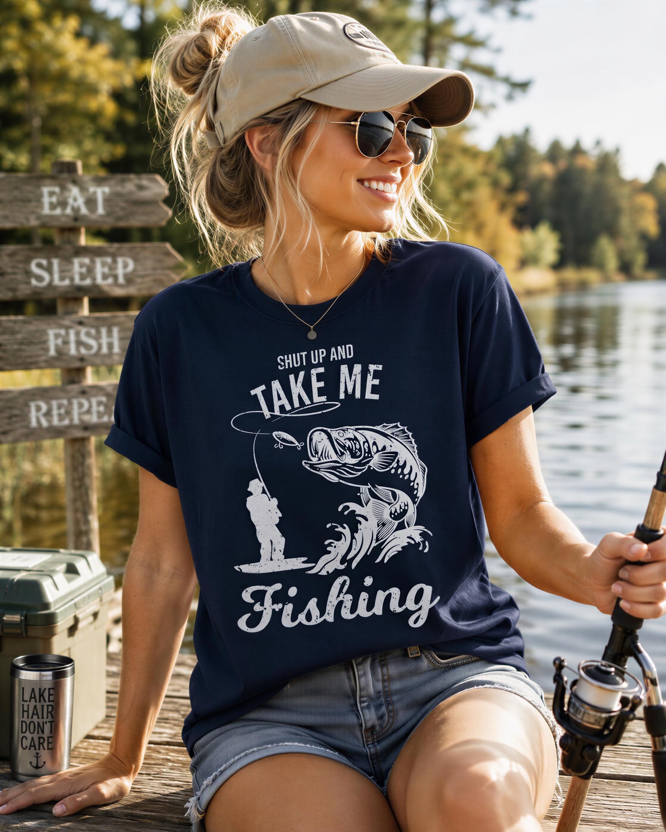 A woman wearing a blue t-shirt with a fishing graphic and the text "Take me fishing" is sitting on a dock near a lake, holding a fishing rod and smiling.