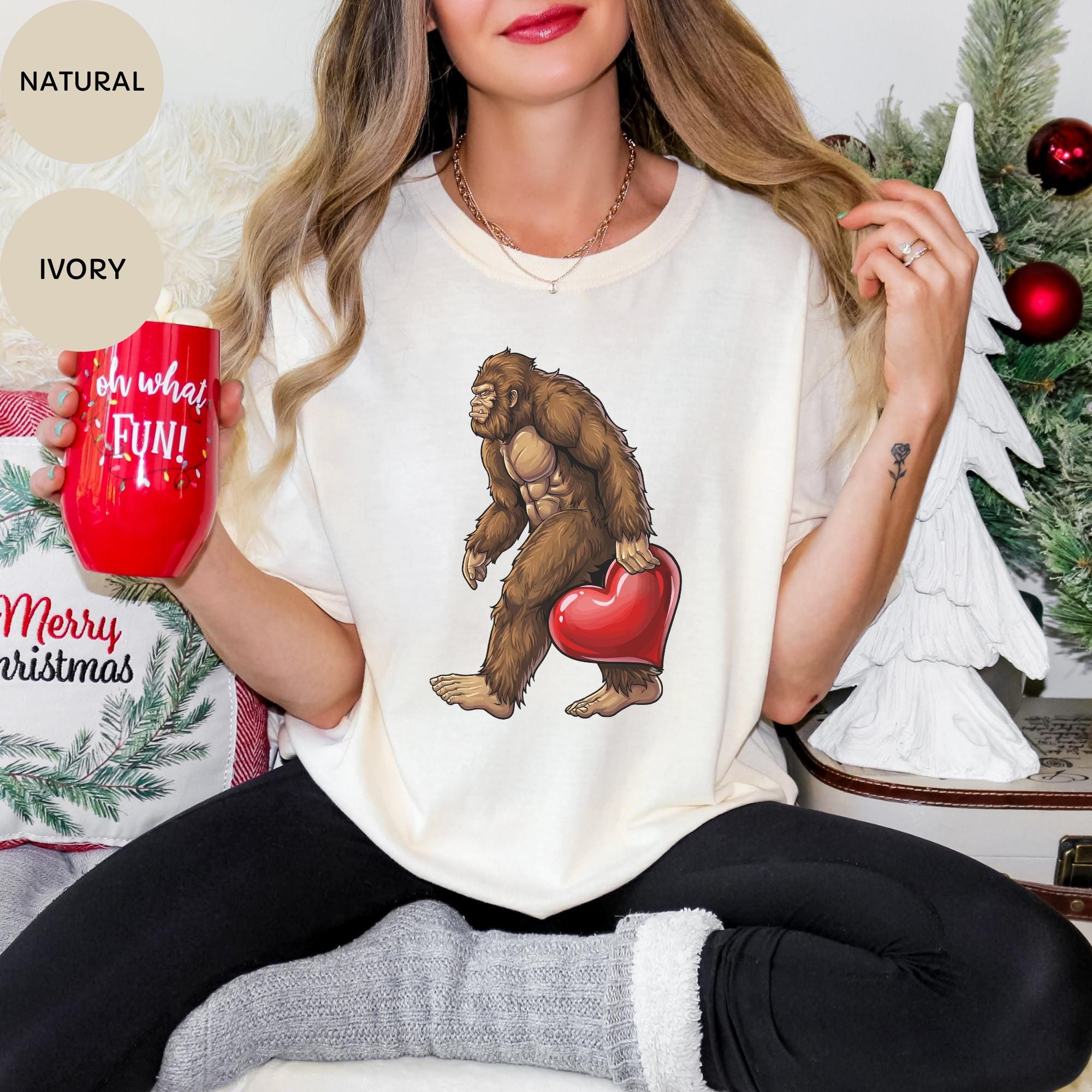 A woman wearing a white t-shirt with a graphic of a brown gorilla holding a red heart, sitting in front of a Christmas tree.