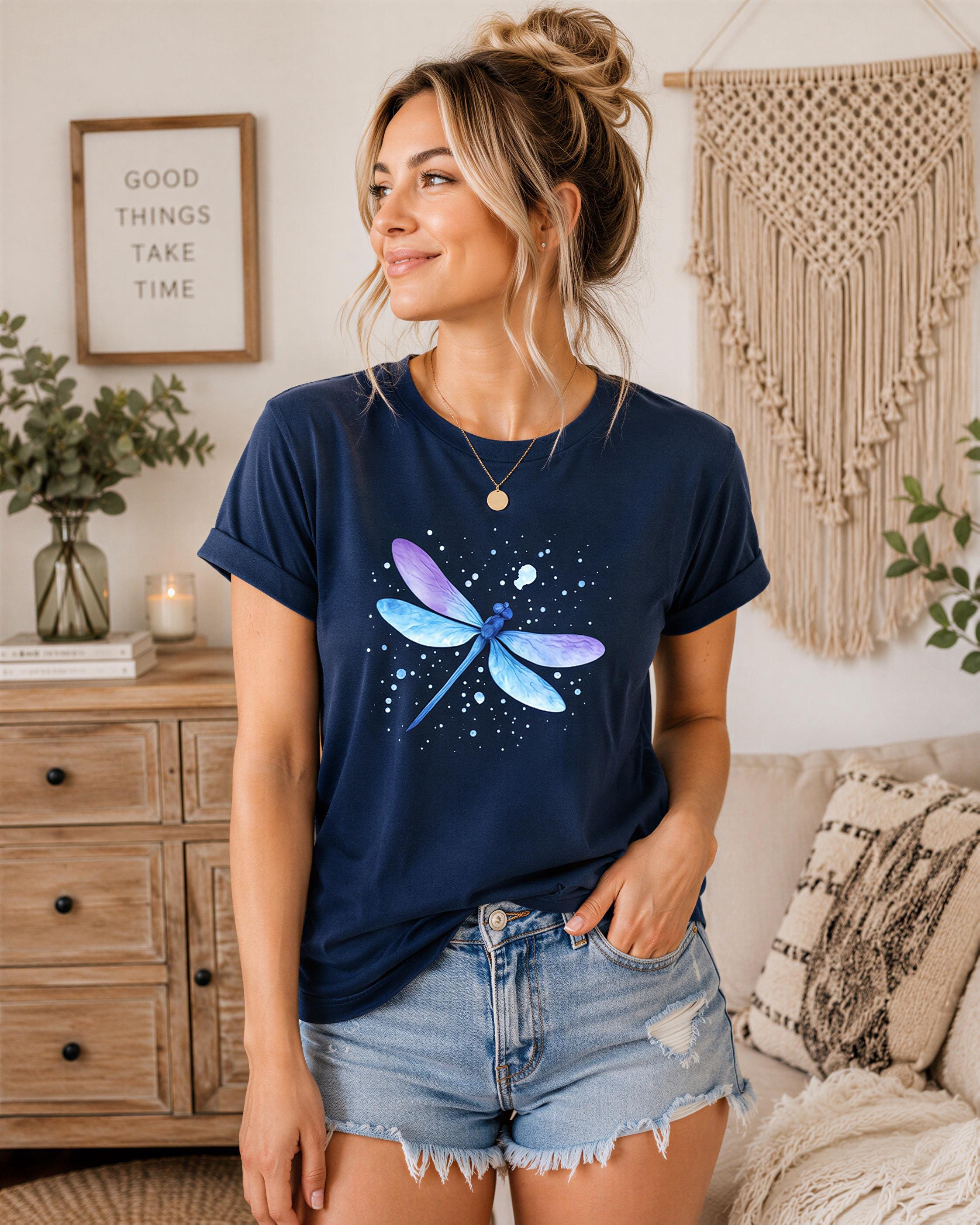 A woman wearing a navy blue t-shirt with a blue and purple dragonfly graphic stands in a room with a wooden dresser, a woven wall hanging, and a potted plant.