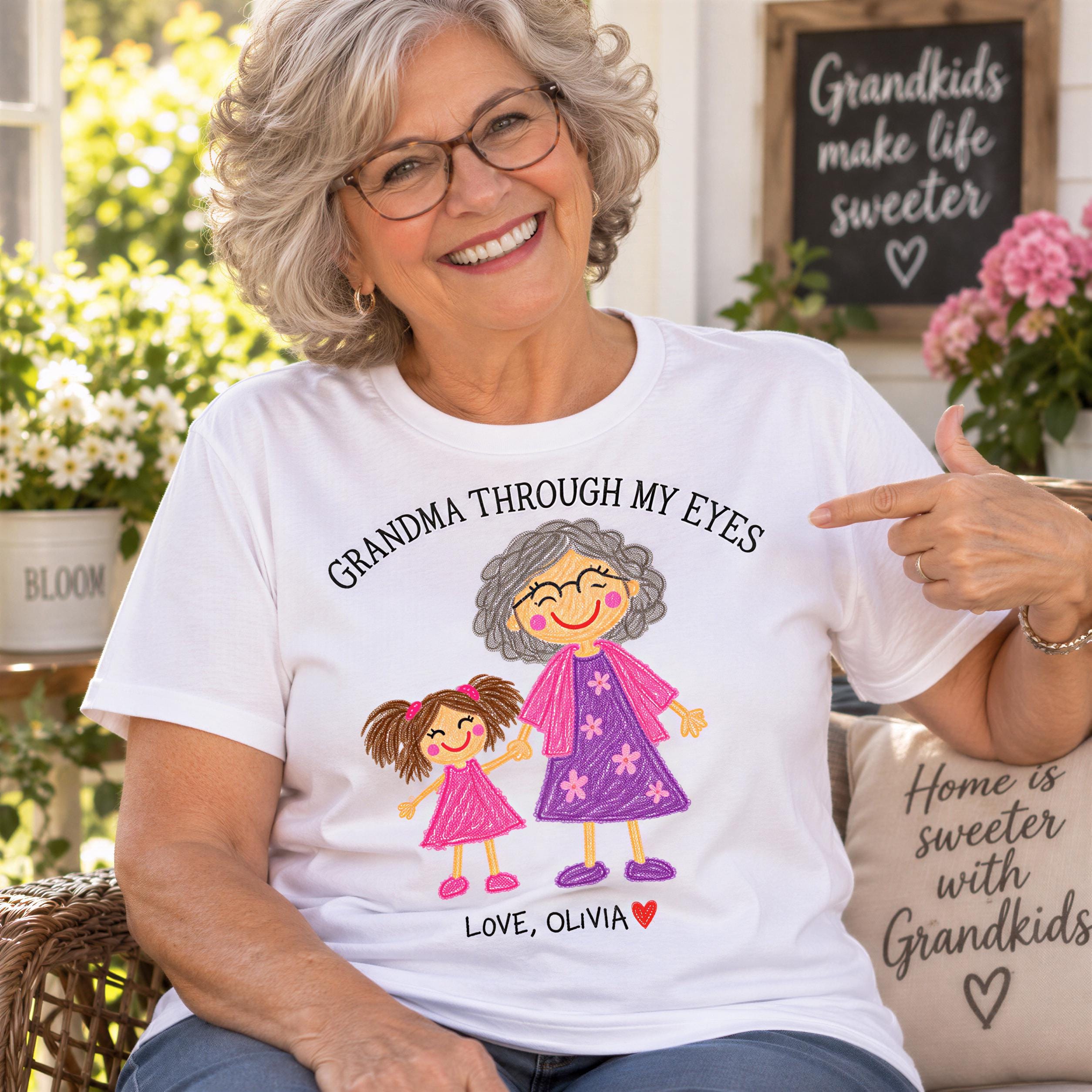A smiling older woman wearing a white t-shirt with a drawing of a young girl and the text "GRANDMA THROUGH MY EYES" is pointing at the shirt.