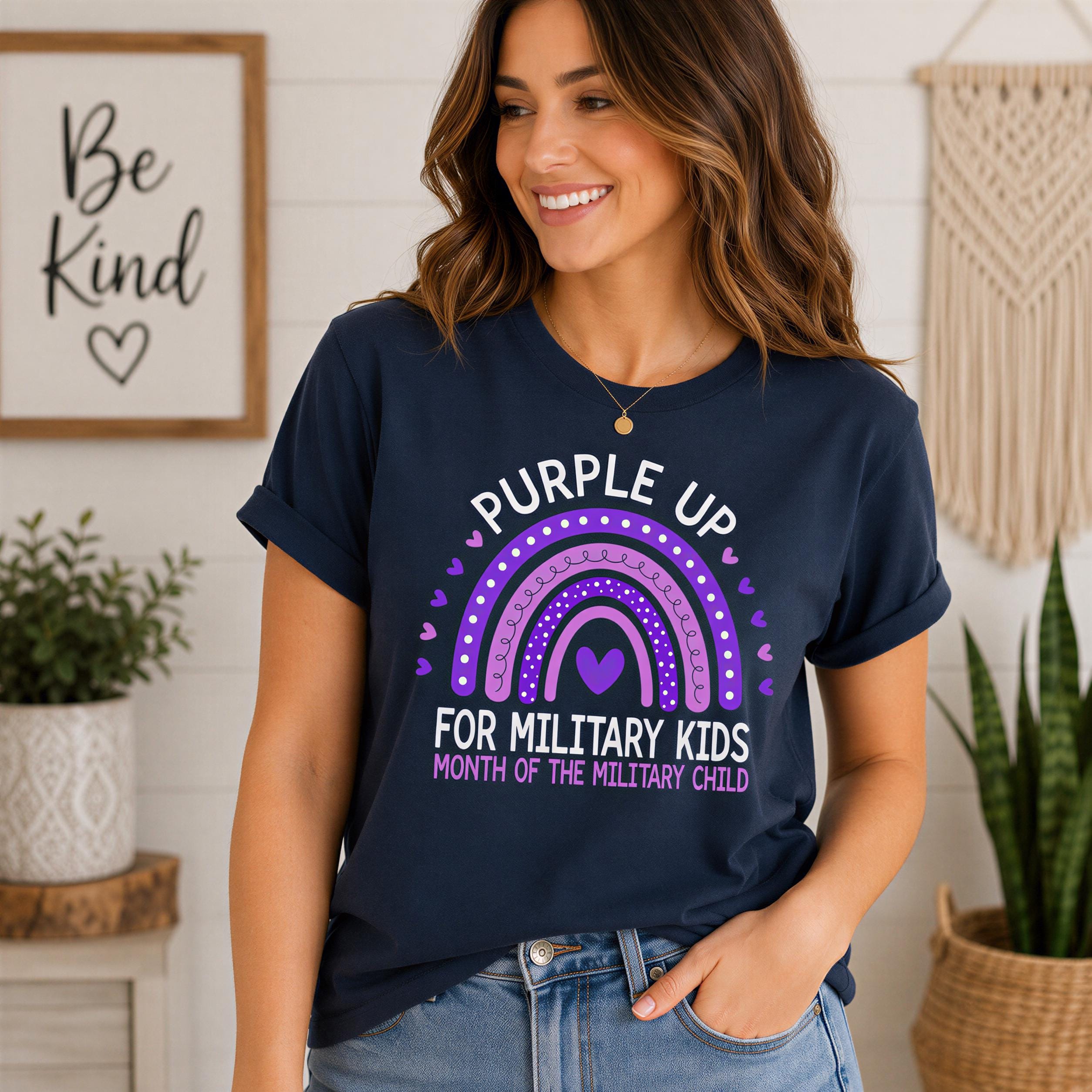 A woman wearing a navy blue t-shirt with a graphic design and text, smiling and standing in front of a wall with a framed quote and a woven decoration.