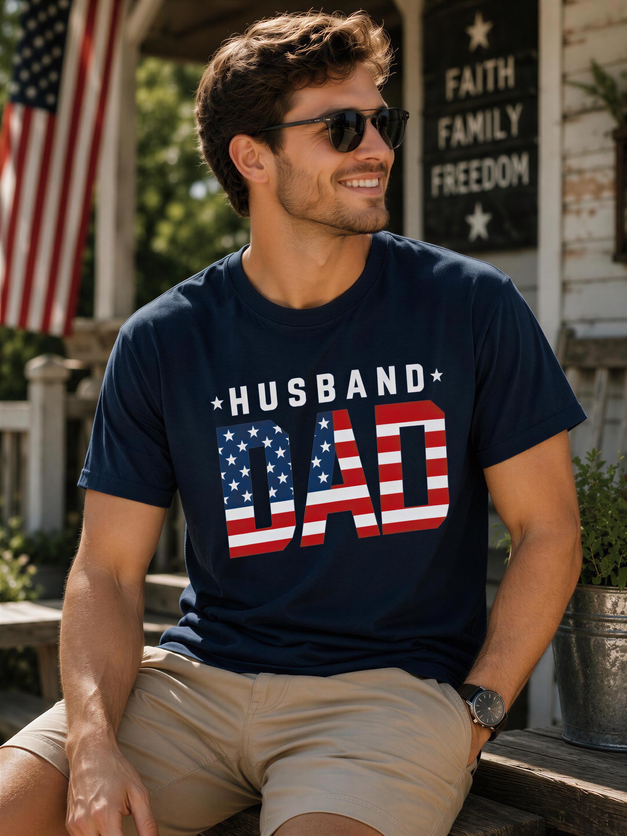 A man wearing a navy blue t-shirt with the text "HUSBAND DAD" and an American flag design, smiling and sitting on a bench.