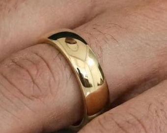 Puede incluir: Un anillo de banda de oro pulido con una superficie lisa y redondeada y un acabado de alto brillo. El anillo presenta un diseño circular simple y continuo sin grabados ni piedras, reflejando la luz en su superficie metálica dorada.