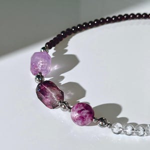 May include: A beaded necklace featuring a strand of small dark purple beads, two faceted translucent purple stones, one round mottled magenta bead, and a series of clear spherical beads, all separated by small metallic silver-toned spacer beads.