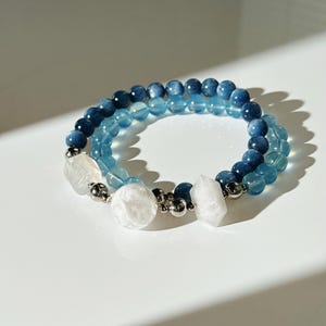 May include: A double-strand beaded bracelet featuring deep blue and light blue round beads. The design includes two clear, faceted hexagonal beads and one large, textured white spherical bead, all accented with small silver-toned metal spacers.
