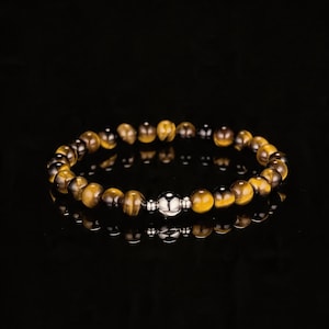 Puede incluir: Una pulsera de cuentas con piedras ojo de tigre en tonos marrón y dorado. La pulsera tiene una cuenta central de metal plateado. Se muestra sobre una superficie negra reflectante.