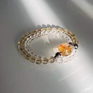 May include: A multi-strand beaded bracelet featuring translucent smoky quartz beads, clear quartz beads, and a single large, spherical orange citrine bead accented by two small, dark metallic spacer beads, arranged in a circular, layered design.