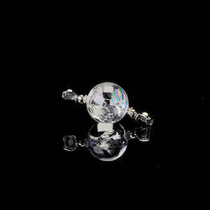 May include: A clear, spherical crystal pendant with iridescent rainbow reflections. The pendant is connected to two silver-colored, rectangular end pieces. The object is set against a black background, with its reflection visible below.