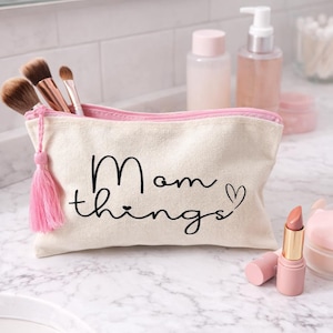 Könnte beinhalten: Eine beigefarbene Kosmetiktasche aus Canvas mit einem rosa Reißverschluss und Quaste, beschriftet mit "Mom things" in schwarzer Schrift. Make-up-Pinsel und Lippenstift sind sichtbar.