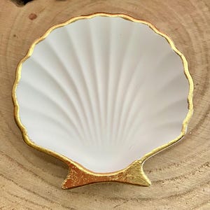 Könnte beinhalten: Eine weiße, muschelförmige Schmuckschale mit goldfarbenem Rand und Boden. Die Schale hat ein strukturiertes, fächerartiges Muster auf der Innenseite. Die Schale ist wahrscheinlich aus Keramik oder Porzellan und dient zur Aufbewahrung kleiner Gegenstände.