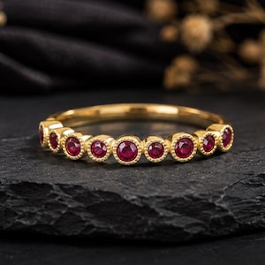 Puede incluir: Un anillo de oro con una hilera de nueve rubíes rojos redondos engastados en bisel. Cada rubí está enmarcado por un delicado borde de metal con cuentas, creando un diseño de banda continua. El anillo está hecho de oro amarillo pulido.