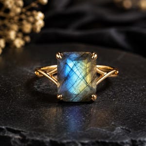 May include: A gold ring featuring a rectangular, faceted labradorite gemstone with blue and yellow iridescence. The stone is held in a four-prong setting on a thin, polished gold band that splits into a V-shape where it meets the gemstone.