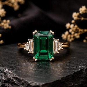 Op de afbeelding: Een gouden ring met een grote smaragdgeslepen groene edelsteen in het midden, geflankeerd door twee kleinere baguettegeslepen heldere stenen en twee kleine ronde heldere stenen aan elke kant, gezet in een gepolijste gouden ringband.
