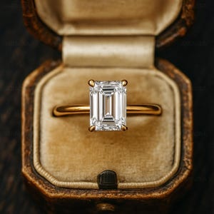 Puede incluir: Un anillo de oro con un diamante de talla esmeralda, engastado en un engaste clásico de cuatro garras. El anillo se presenta en un joyero de terciopelo marrón de estilo vintage.