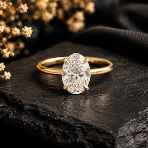 Puede incluir: Un anillo de oro con una piedra preciosa transparente de corte ovalado sujeta por cuatro garras. La banda es lisa y pulida, mostrando un diseño clásico con una piedra central que refleja la luz en su superficie facetada.