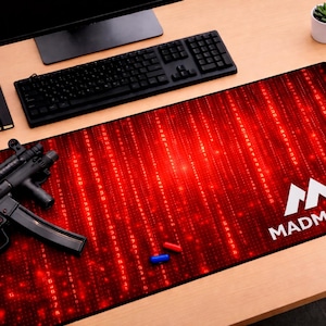 Könnte beinhalten: Rotes und schwarzes MADMATCO Gaming-Mauspad mit Matrix-Design. Eine schwarze Tastatur, ein Monitor und ein Notizbuch befinden sich auf dem Holztisch. Eine schwarze MP5-Maschinenpistole und zwei kleine Kapseln liegen ebenfalls auf dem Tisch.