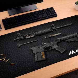 Puede incluir: Alfombrilla de escritorio negra con armas de fuego desmontadas y munición. La alfombrilla presenta el logotipo "MADMATCO" en blanco. También se ven un teclado, un monitor y un cuaderno negros.