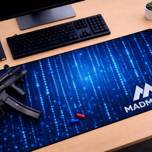 May include: A black computer keyboard, monitor, and mousepad with a blue matrix design and the text "MADMATCO". A black notebook, a small potted plant, and a black firearm are also on the desk.