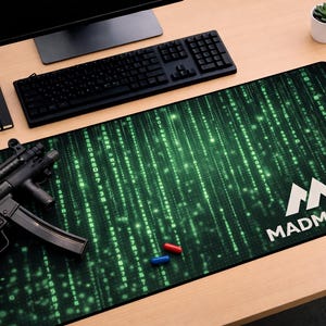 May include: A large black and green desk mat with a digital rain pattern and the white text "MADMATCO". A black keyboard, notebook, and a small succulent plant are also on the desk. A black firearm and two colored pills are on the mat.