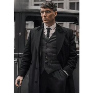 Può includere: Un uomo con un cappotto nero, un gilet e un completo, con una camicia bianca con colletto e una cravatta fantasia. Indossa occhiali rotondi e ha una catena per orologio da taschino. Un'auto d'epoca è sullo sfondo.