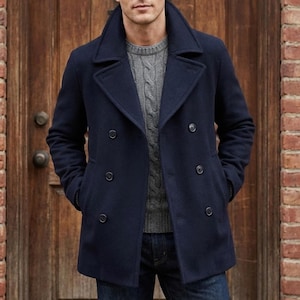 May include: A navy blue double-breasted pea coat with six dark buttons and a wide collar. The coat is worn over a gray sweater and blue jeans. The background features a wooden door and a brick wall.