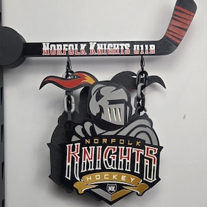 May include: A wall-mounted sign featuring a black hockey stick with red tape, labelled Norfolk Knights U11B. Hanging from the stick by black chains is a multi-layered emblem displaying a knight helmet, flames, and the text Norfolk Knights Hockey NK.