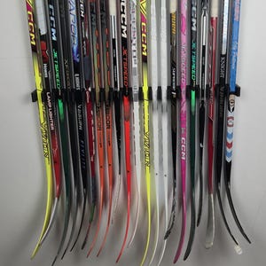 May include: A collection of eighteen ice hockey sticks mounted vertically on a wall rack. The sticks feature various brand names including CCM, Bauer, and Vapor, with diverse colour schemes such as yellow, black, red, white, pink, and blue.