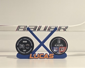 Könnte beinhalten: Ein Hockey-Display mit einer blau-orangen Basis mit dem Namen "LUCAS". Zwei Hockey-Pucks und ein Hockeyschläger sind sichtbar. Der Hockeyschläger trägt den Markennamen "BAUER". Die Pucks haben Text und Logos.