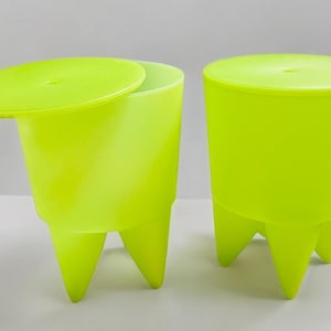Può includere: Due sgabelli in plastica verde neon con tre gambe ciascuno. Uno sgabello ha un coperchio parzialmente aperto. Gli sgabelli sono cilindrici con una parte superiore leggermente svasata. Sono in plastica liscia e lucida.