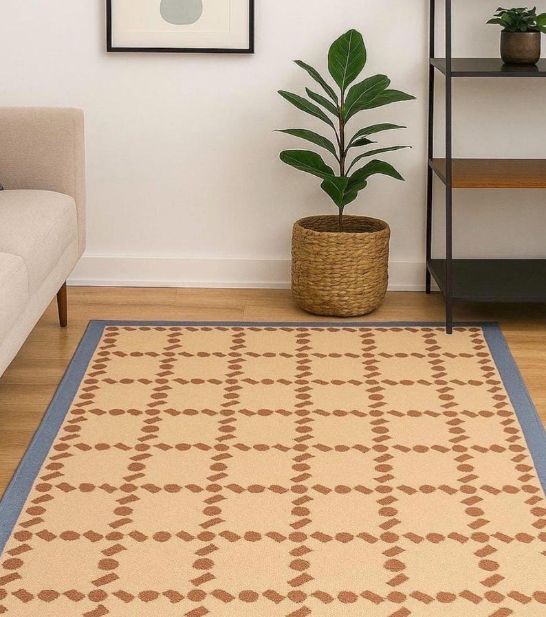 Puede incluir: Alfombra rectangular con estampado geom&eacute;trico beige y marr&oacute;n y borde azul. El dise&ntilde;o de la alfombra consiste en formas marrones repetidas sobre un fondo beige. Est&aacute; colocada sobre un suelo de madera.