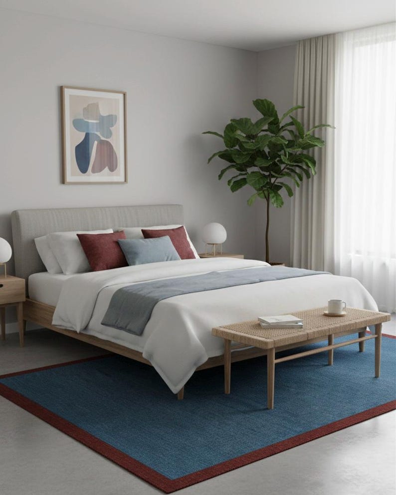 Puede incluir: Interior de dormitorio con una cama con ropa de cama blanca, cojines decorativos y un camino de cama azul. Un banco tejido se encuentra al pie de la cama. Una alfombra azul con borde rojo y una obra de arte abstracta enmarcada.