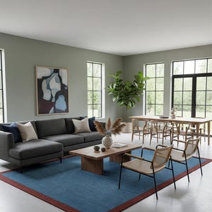Puede incluir: Un sal&oacute;n moderno con un sof&aacute; seccional gris, una mesa de centro de madera y una alfombra azul con borde rojo. Dos sillas de mimbre frente al sof&aacute;. Una mesa de comedor y sillas al fondo, junto a grandes ventanales y una pintura abstracta.