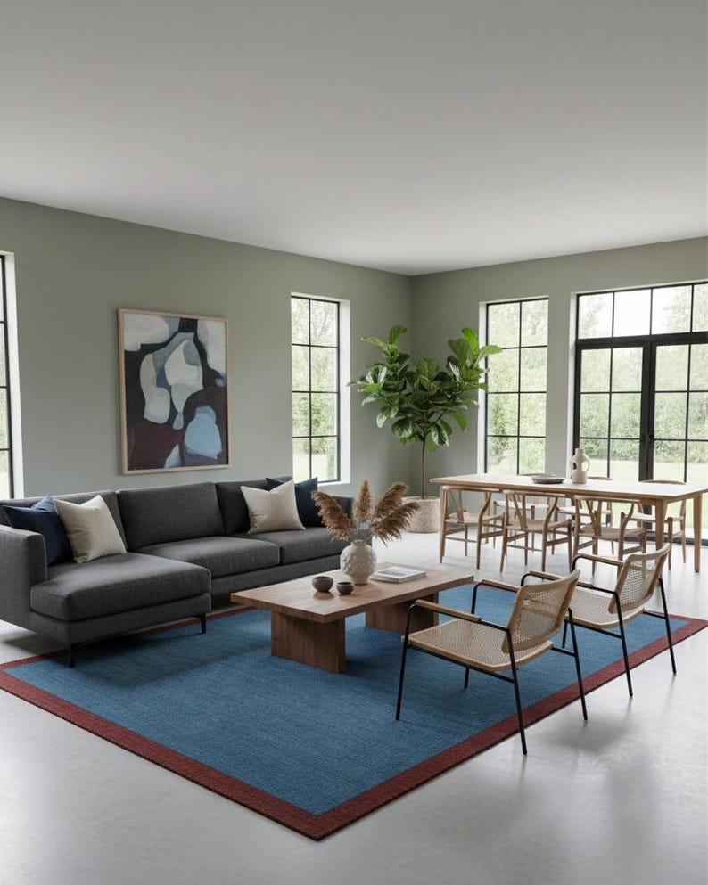 Puede incluir: Un sal&oacute;n moderno con un sof&aacute; seccional gris, una mesa de centro de madera y una alfombra azul con borde rojo. Dos sillas de mimbre frente al sof&aacute;. Una mesa de comedor y sillas al fondo, junto a grandes ventanales y una pintura abstracta.