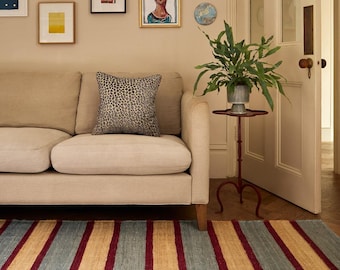 May include: A beige sofa with a leopard print pillow sits in front of a wall adorned with framed artwork. A red side table holds a potted plant. A striped rug in blue, gold, and red covers the floor.