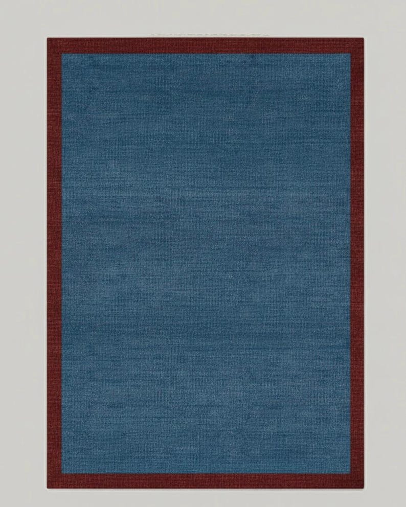 Puede incluir: Una alfombra rectangular con un centro azul y un borde rojo oscuro. La alfombra tiene una apariencia texturizada, lo que sugiere una construcci&oacute;n tejida o de tela. La paleta de colores es simple y elegante, adecuada para varios estilos de dise&ntilde;o de interiores.