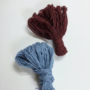 Puede incluir: Dos ovillos de lana en colores contrastantes. Uno es de color burdeos intenso, el otro azul apagado. Cada ovillo est&aacute; atado en la base, con los hilos extendidos. Ideal para proyectos de tejido o ganchillo.