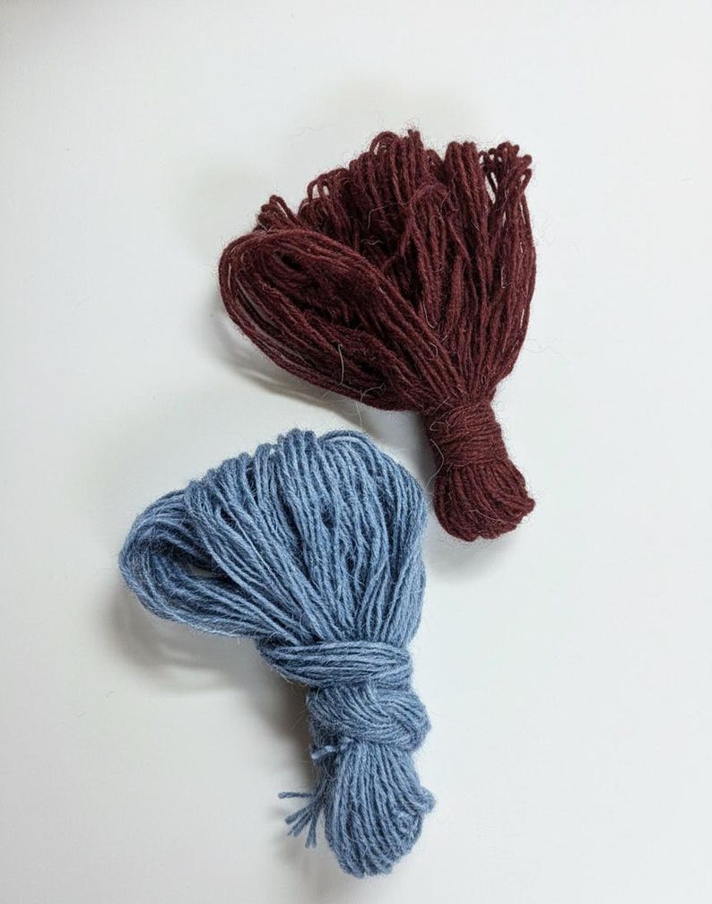 Puede incluir: Dos ovillos de lana en colores contrastantes. Uno es de color burdeos intenso, el otro azul apagado. Cada ovillo est&aacute; atado en la base, con los hilos extendidos. Ideal para proyectos de tejido o ganchillo.