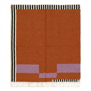 May include: A woven rug with a geometric design. The rug features a large rectangular field of rust-orange, with two lavender rectangles and a border of black and white stripes. The rug has a fringe on the bottom edge.