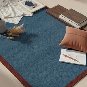 Puede incluir: Una alfombra rectangular azul con un borde rojo oscuro. La alfombra est&aacute; rodeada de elementos de decoraci&oacute;n del hogar, incluyendo una impresi&oacute;n art&iacute;stica abstracta enmarcada, un coj&iacute;n marr&oacute;n y una pila de libros. Tambi&eacute;n hay un jarr&oacute;n y plantas secas.
