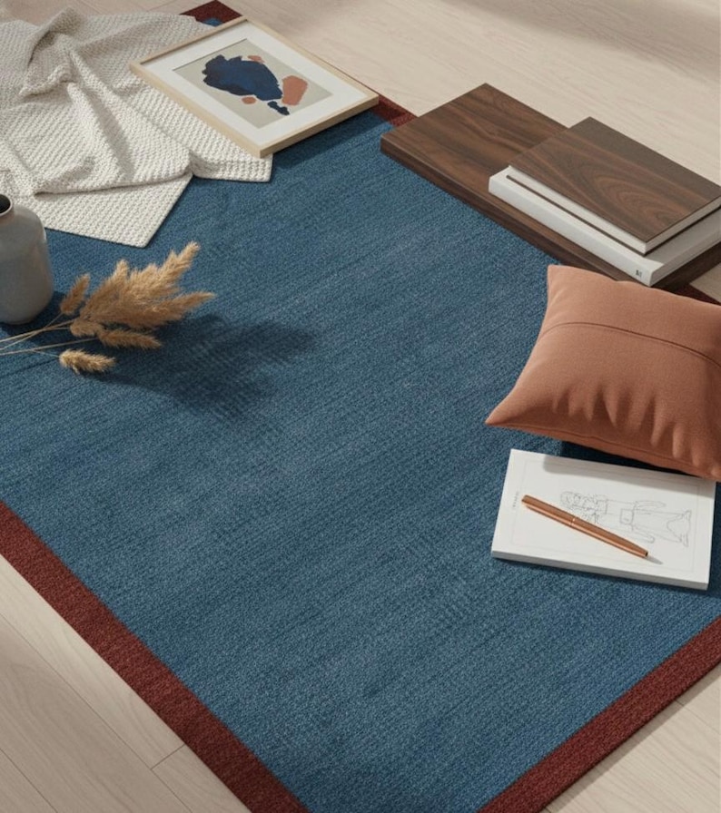 Puede incluir: Una alfombra rectangular azul con un borde rojo oscuro. La alfombra est&aacute; rodeada de elementos de decoraci&oacute;n del hogar, incluyendo una impresi&oacute;n art&iacute;stica abstracta enmarcada, un coj&iacute;n marr&oacute;n y una pila de libros. Tambi&eacute;n hay un jarr&oacute;n y plantas secas.