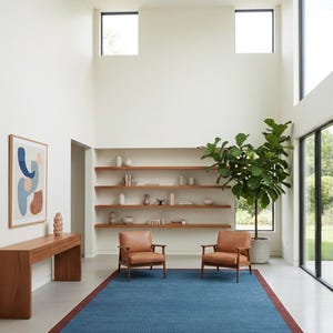 Puede incluir: Un sal&oacute;n moderno con una alfombra azul con borde rojo, dos sillones de cuero marr&oacute;n y una consola de madera. La habitaci&oacute;n tiene una gran planta en maceta, estanter&iacute;as y grandes ventanas.