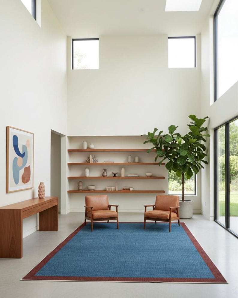 Puede incluir: Un sal&oacute;n moderno con una alfombra azul con borde rojo, dos sillones de cuero marr&oacute;n y una consola de madera. La habitaci&oacute;n tiene una gran planta en maceta, estanter&iacute;as y grandes ventanas.